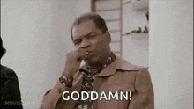 a man in a brown jacket is smoking a cigarette and saying goddamn .