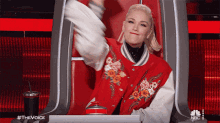 a woman wearing a red jacket with flowers on it is sitting in a chair with a nbc logo on the bottom