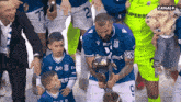 a man in a blue jersey with the number 2 on it holds a trophy in front of a group of soccer players