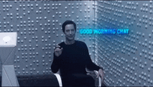 a man in a black shirt is sitting in front of a wall that says good morning chat .