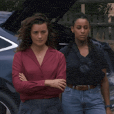 two women standing next to each other with their arms crossed in front of a car