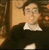a man wearing glasses is smiling in front of a wall with a clock on it