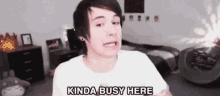 a young man is sitting in a bedroom with the words kinda busy here written on his shirt .