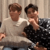 two men are sitting next to each other on a bed . one of the men is holding a cell phone .