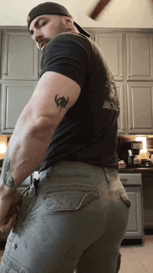 a man with a tattoo on his arm is standing in a kitchen wearing cargo pants and a hat .