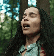 a woman with dreadlocks is standing in the woods with her eyes closed and her mouth open .