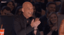 a bald man is sitting in front of a crowd clapping and smiling .