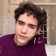 a young man with curly hair is looking at the camera with mathematical equations drawn on the wall behind him