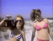 two women in bikinis are walking on the beach . one is holding a camera .