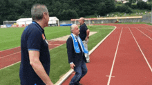 a man with a blue scarf around his neck walks on a track in a stadium