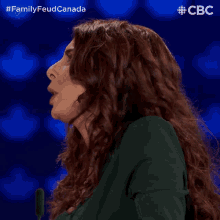 a woman with long red hair stands in front of a microphone with the words family feud canada on the bottom right