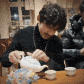 a man in a black turtleneck is pouring tea into a cup