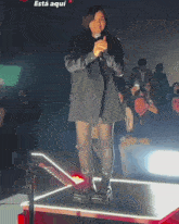 a woman stands on a stage with a microphone in front of a crowd and the word está aqui is on the bottom right