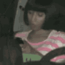 a woman in a pink and white striped shirt is sitting in a car using a cell phone .