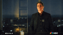 a man in a suit stands in front of a window with nbc written on the bottom right