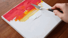 a person is using a spatula to spread red and yellow paint on a white canvas