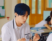 a boy with blue hair is sitting at a desk in a classroom with a ring on his finger