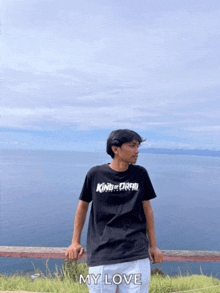 a man wearing a black shirt that says king of drag stands in front of the ocean