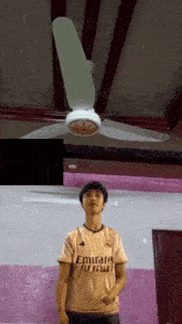 a boy wearing an emirates jersey stands in front of a fan