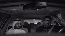 a man with a beard wearing a white shirt and a head scarf sits in a car with his eyes closed