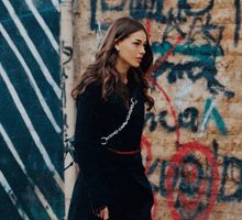 a woman in a black dress stands in front of a brick wall with graffiti on it