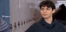 a young man in a blue sweater is standing in front of a row of lockers in a locker room .