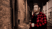 a man wearing glasses and a red plaid shirt stands in an alleyway