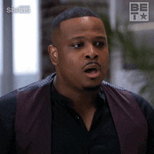 a man wearing a black shirt and a burgundy vest is standing in front of a sign that says sistas on it