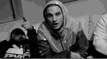 a man in a hooded jacket is giving a thumbs up sign while sitting on a couch .