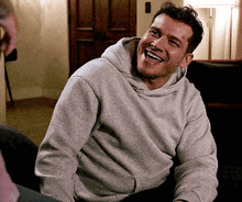 a man in a grey hoodie is sitting on a couch laughing .