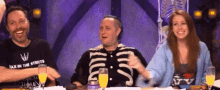 a group of people are sitting at a table with drinks . one of the men is wearing a skeleton shirt .
