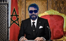 a man in a suit and tie is sitting at a table in front of a flag .