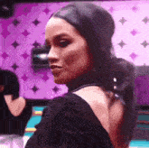 a woman in a black dress is standing in front of a purple wall and looking at the camera .
