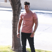 a man wearing a pink t-shirt and black jeans is standing in front of a tree