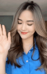 a woman with long hair is wearing a blue shirt and waving at the camera .