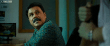 a man with a mustache is sitting at a desk in front of a computer screen .
