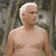 a shirtless man with white hair is standing in front of a house and trees .