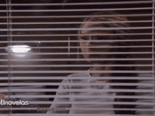 a woman is looking through a window with blinds and the words tinovelas on the bottom right