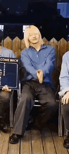 a man with blonde hair is sitting in a chair with his arms outstretched while holding a sign that says come back .
