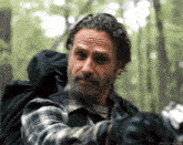 a man with a beard wearing a plaid shirt is standing in the woods with a backpack on his back