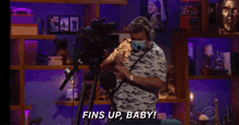 a man wearing a mask stands in front of a camera with the words fins up baby behind him
