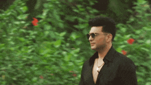 a man in a black shirt and sunglasses stands in front of a bush