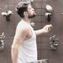a man in a white tank top is standing in front of a wall with plants hanging from it