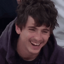 a close up of a young man laughing with his eyes closed and wearing a purple hoodie .