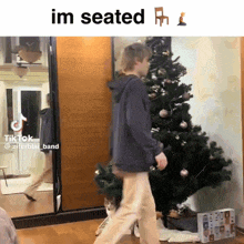 a man is walking in front of a christmas tree with the caption " im seated " above him