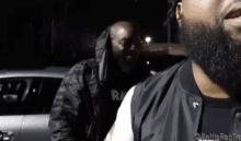 a man with a beard is standing next to another man in front of a car at night .