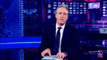 a man in a suit and tie is sitting at a desk with a comedy central logo on the bottom