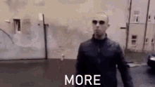 a man in a black jacket and sunglasses is walking down a street with the words `` more '' behind him .