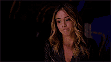 a woman in a leather jacket is sitting in a dark room and looking at the camera .