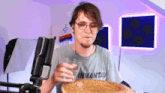 a man wearing glasses holds a glass of water in front of a microphone while eating a pizza